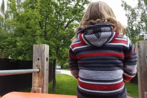 a young child wearing a black and red sweater is sitting a top a playground structure, looking away from the camera