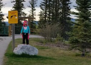 Young girl wearing a pink superhero cape, standing on a large rock outdoors