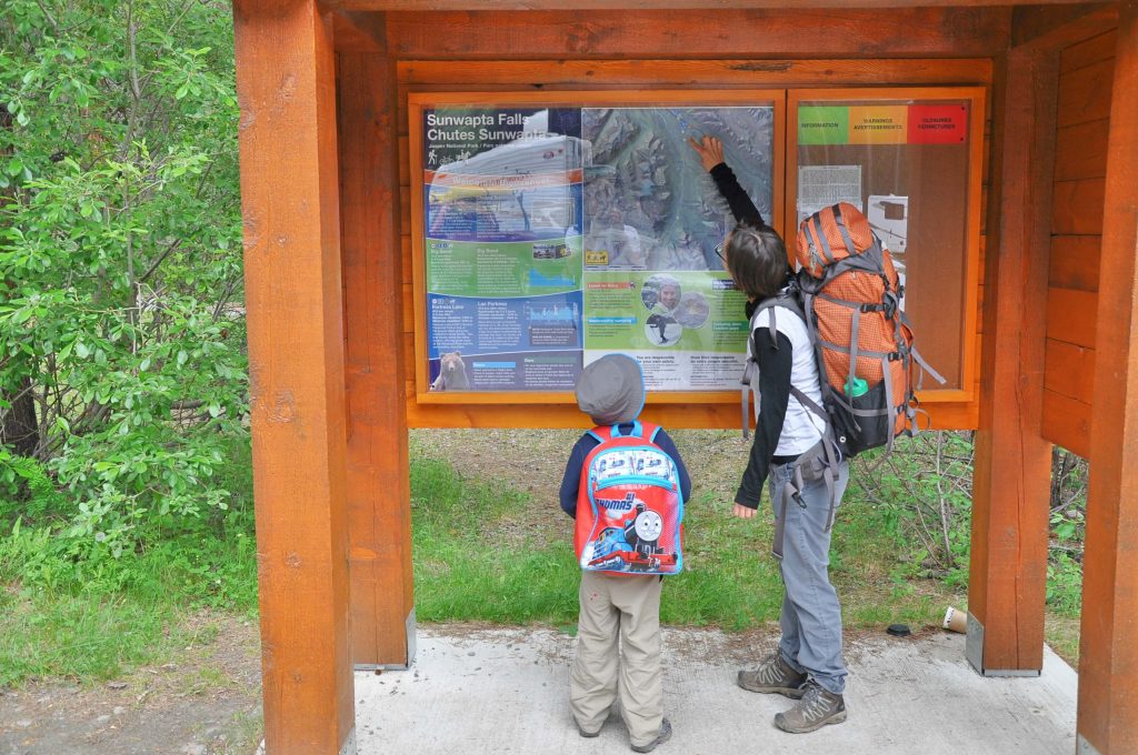 Mom wearing a backpacker's pack is pointing the trail out on a map to her small child, also wearing a small backpack