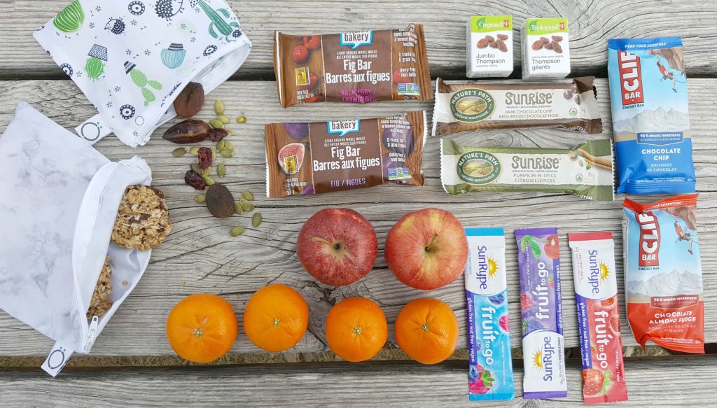 various snack foods like granola bars, fruit, nuts, and dried fruit on a picnic table