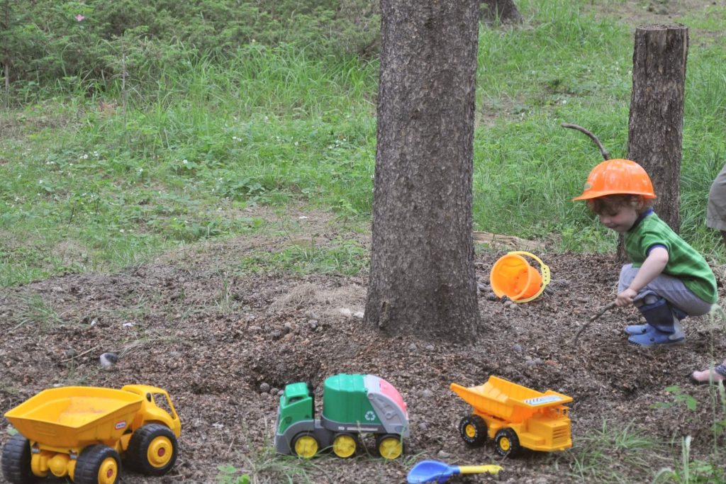Little boy wearing a play hard hat playing with toy trucks and tractors outside in the dirt