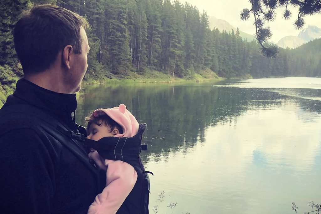 Dad exploring nature with his baby. He is looking out over a mountain lake while baby sleeps in a front baby carrier