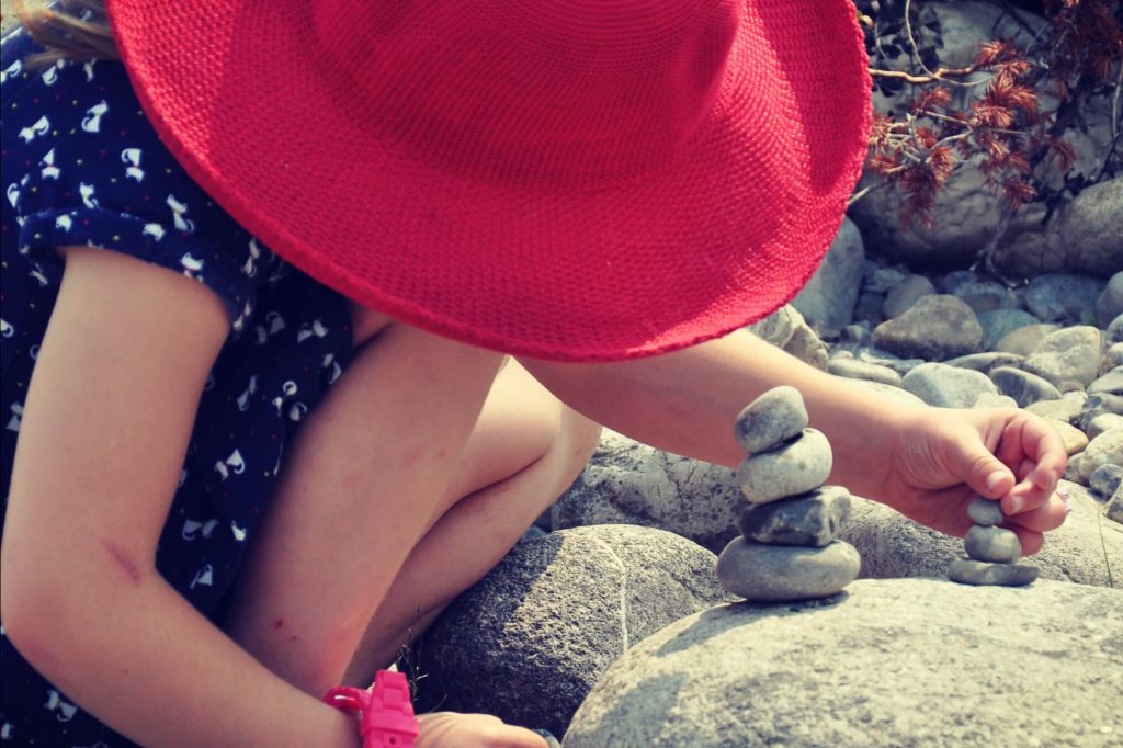 young girl wearing a large red sunhat being creative outdoors by building with stones on the shore