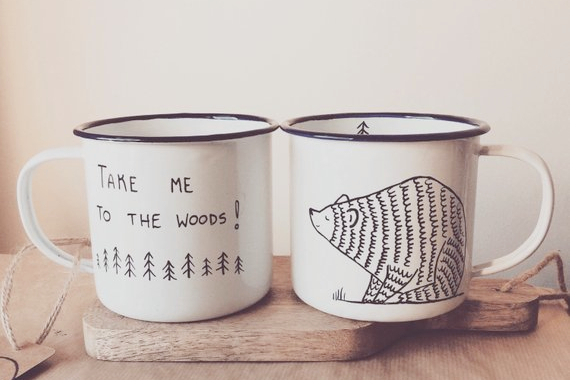 Two white enamel camping mugs on a wooden table. One mug reads take me to the woods. The other is an illustration of a bear.