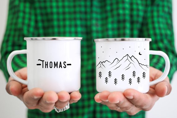 a man with a green plaid shirt holding a personalized enamel mug with mountains and forests