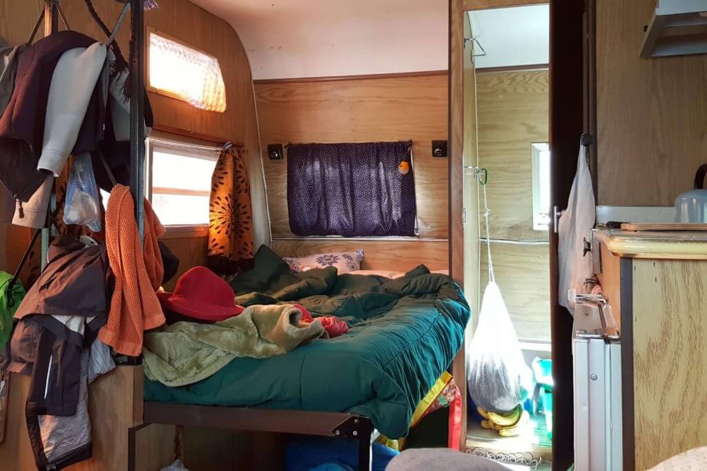 The sleeping area of a small messy trailer, blankets on the bed, clothes strewn around