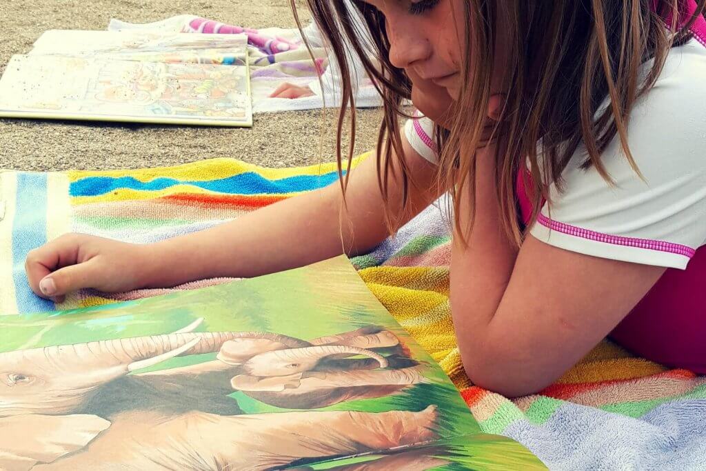 young girl reading a book laying on a towel at the beach