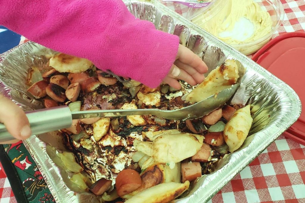a child's hand reaching into a foil dish of perogies and sausage
