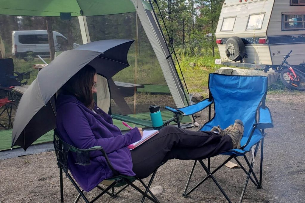 A woman sitting in a camping chair using an umbrella to stay dry at the campground