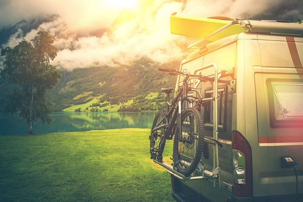 An RV with bikes attached to the back with green fields and mountains in the background
