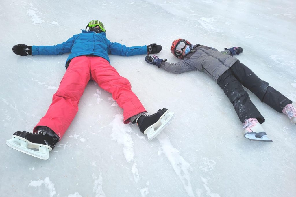 2 kids wearing ice skates wearing winter gear and helmets laying on the frozen lake