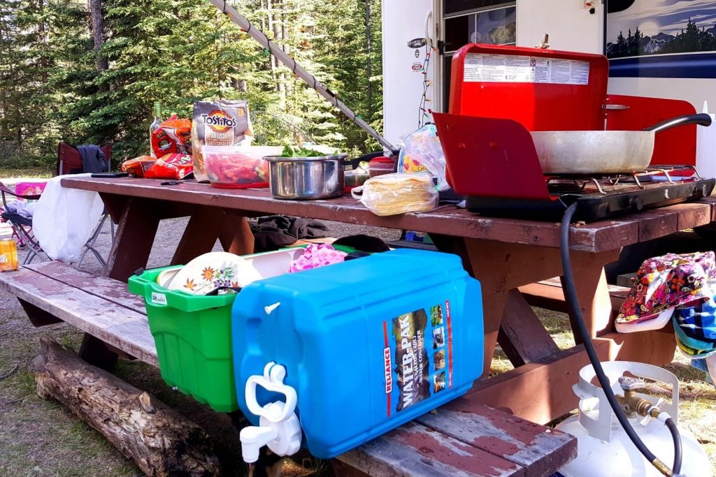 various camp cooking gear on a picnic table - a camp stove, water jug, dishes, pots, food