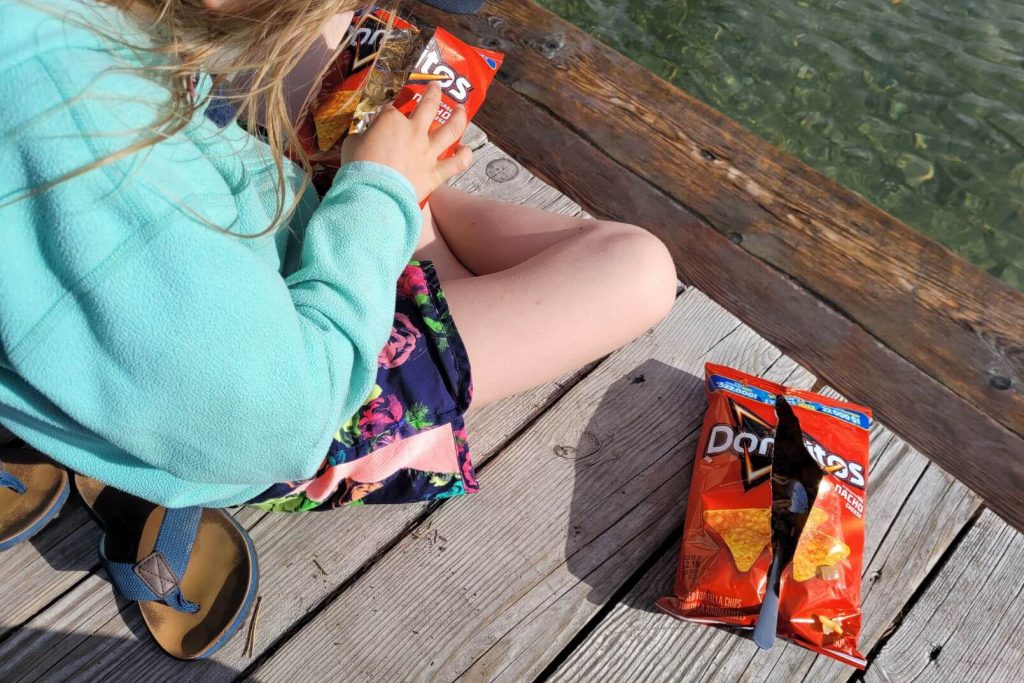 a girl sitting on a dock eating taco bags, a taco in a doritos bag beside her on the dock