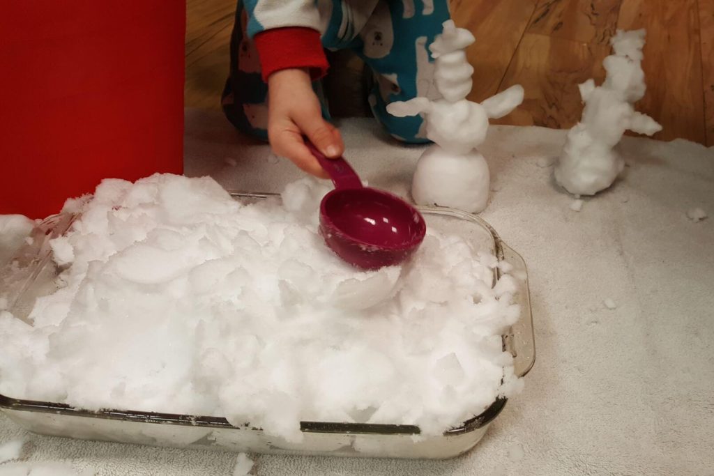 small child's hand holding a measuring spoon, playing with a tub of snow