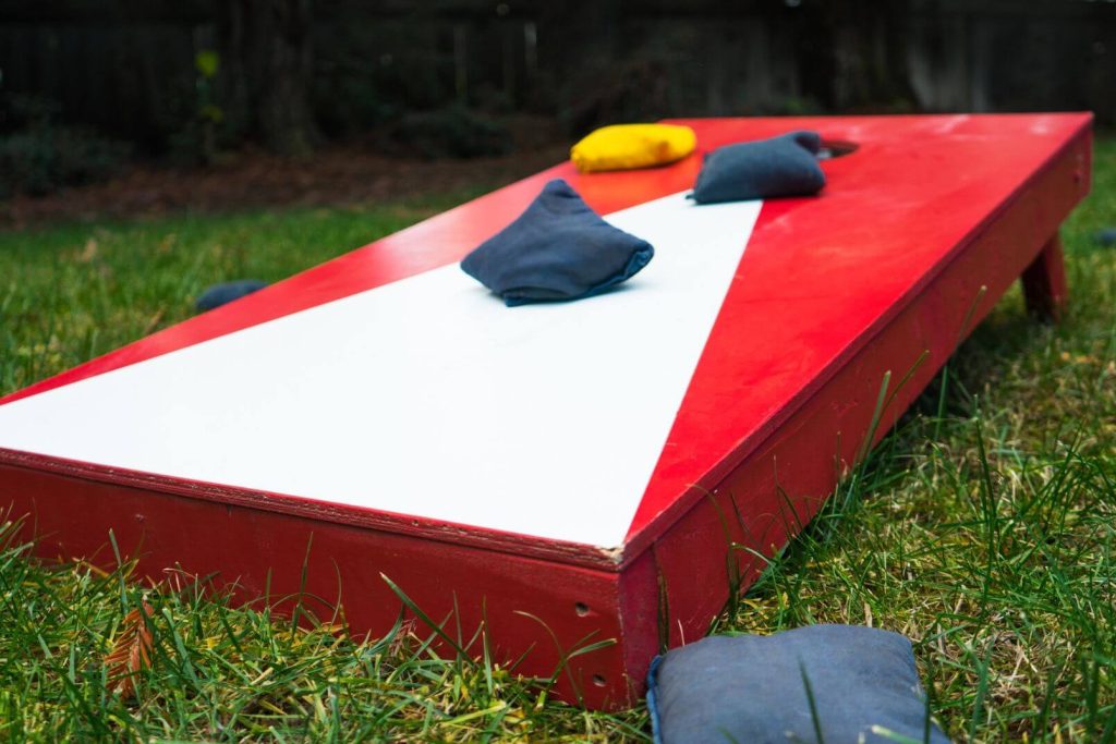 Red and White Corn hole board with bean bags on top and on the grass beside